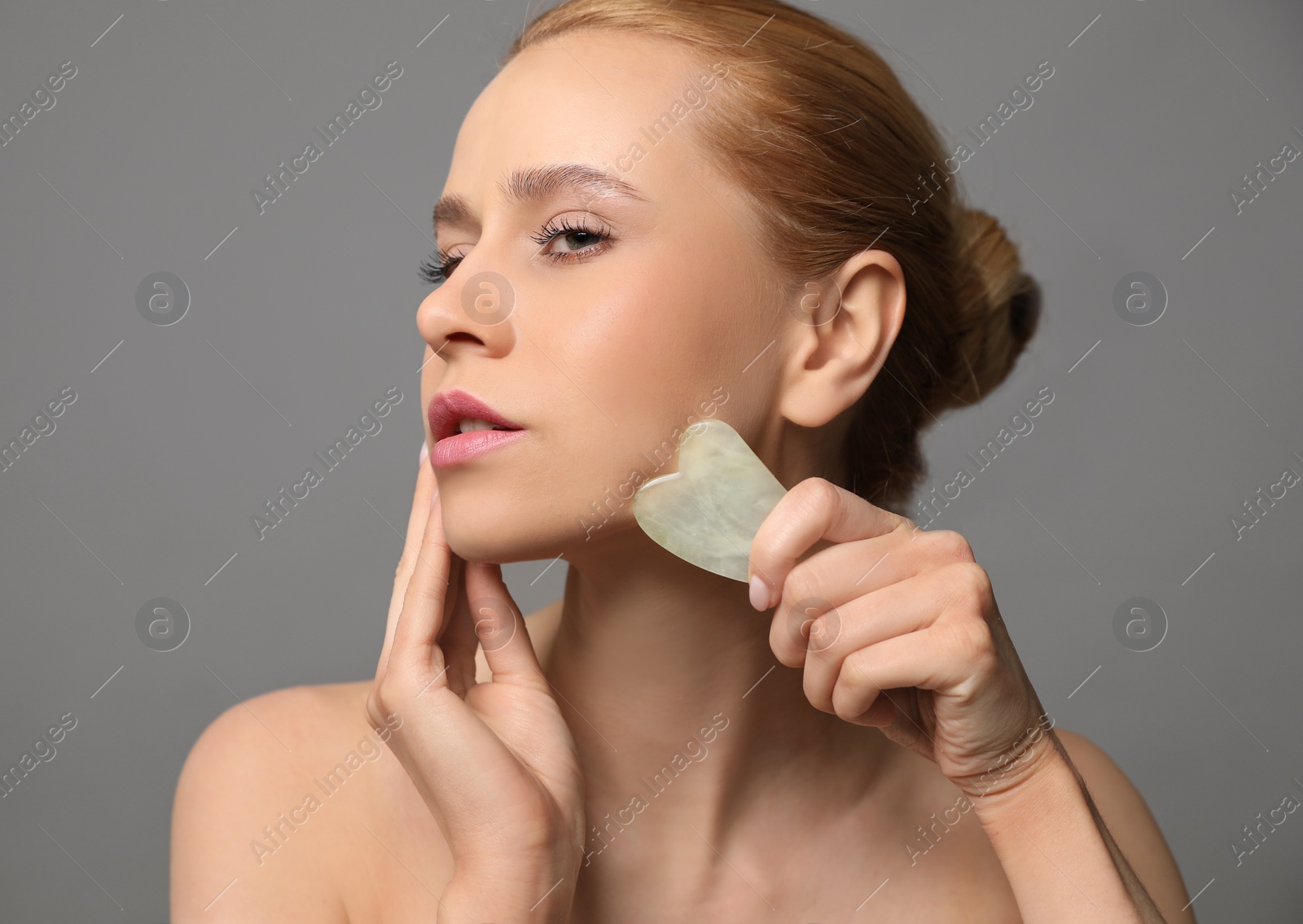 Photo of Beautiful young woman doing facial massage with gua sha tool on grey background, closeup