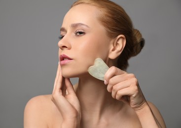 Beautiful young woman doing facial massage with gua sha tool on grey background, closeup