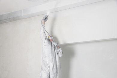 Decorator in protective overalls painting ceiling with spray gun indoors