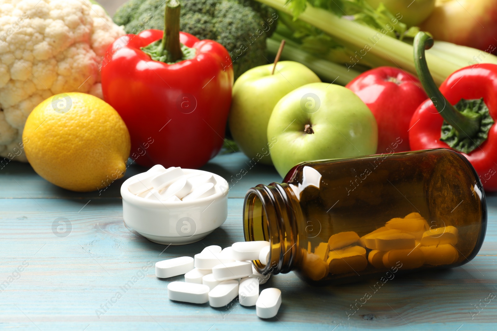 Photo of Dietary supplements. Overturned bottle and bowl with pills near food products on light blue wooden table