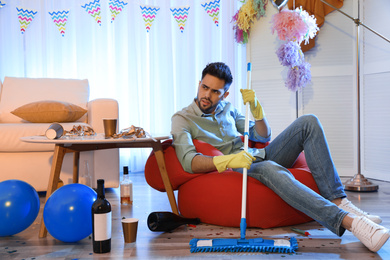 Young man with mop suffering from hangover in messy room after party