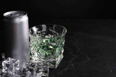 Photo of Energy drink in glass, aluminium can and ice cubes on black table. Space for text