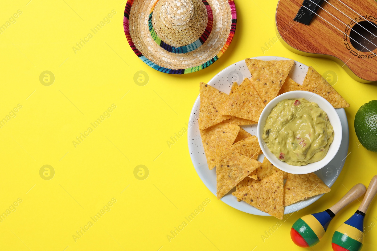 Photo of Mexican sombrero hat, nachos chips, guacamole, maracas and ukulele on yellow background, flat lay. Space for text