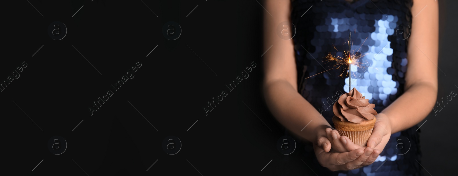 Image of Woman holding birthday cupcake with sparkler on black background, closeup view with space for text. Banner design