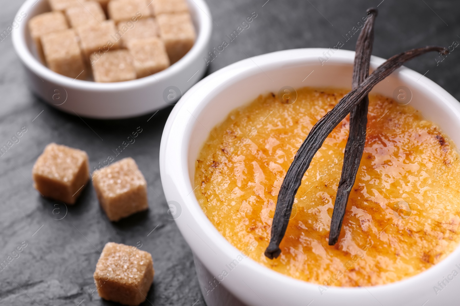 Photo of Delicious creme brulee in bowl, vanilla pods and sugar cubes on dark gray table, closeup