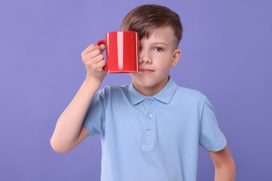 Photo of Cute boy covering eye with red ceramic mug on violet background