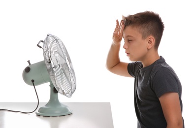 Photo of Little boy suffering from heat in front of fan on white background. Summer season