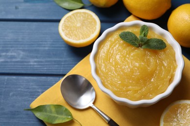 Photo of Delicious lemon curd in bowl, fresh citrus fruits and spoon on blue wooden table, above view
