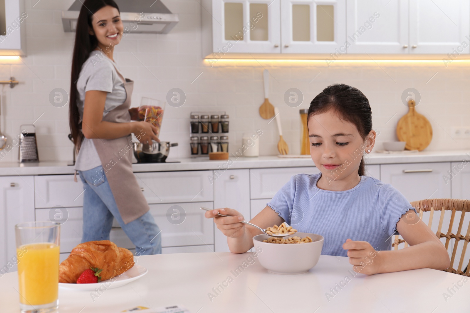 Photo of Little girl having breakfast while her mother cooking food in kitchen. Single parenting