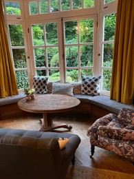 Wooden table near bay window seat with cushions in cozy room. Interior design