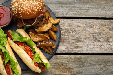 Photo of Tasty burger, hot dogs, potato wedges and sauce on wooden table, top view with space for text. Fast food