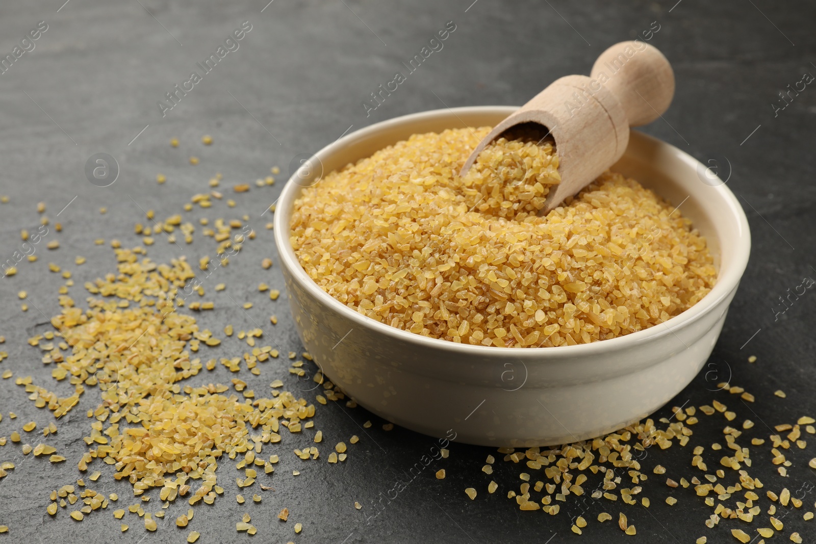 Photo of Bowl and scoop with raw bulgur on dark gray table, closeup