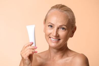 Portrait of beautiful mature woman with perfect skin holding tube of cream on beige background