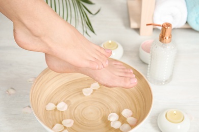 Photo of Woman putting her feet into bowl with water and rose petals indoors, closeup. Spa treatment