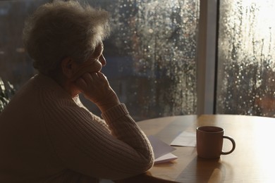 Elderly woman with drink looking out of window indoors on rainy day, space for text. Loneliness concept