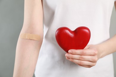 Blood donation concept. Woman with adhesive plaster on arm holding red heart against grey background, closeup