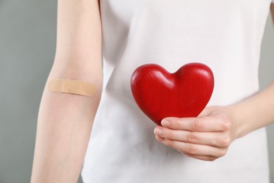 Photo of Blood donation concept. Woman with adhesive plaster on arm holding red heart against grey background, closeup