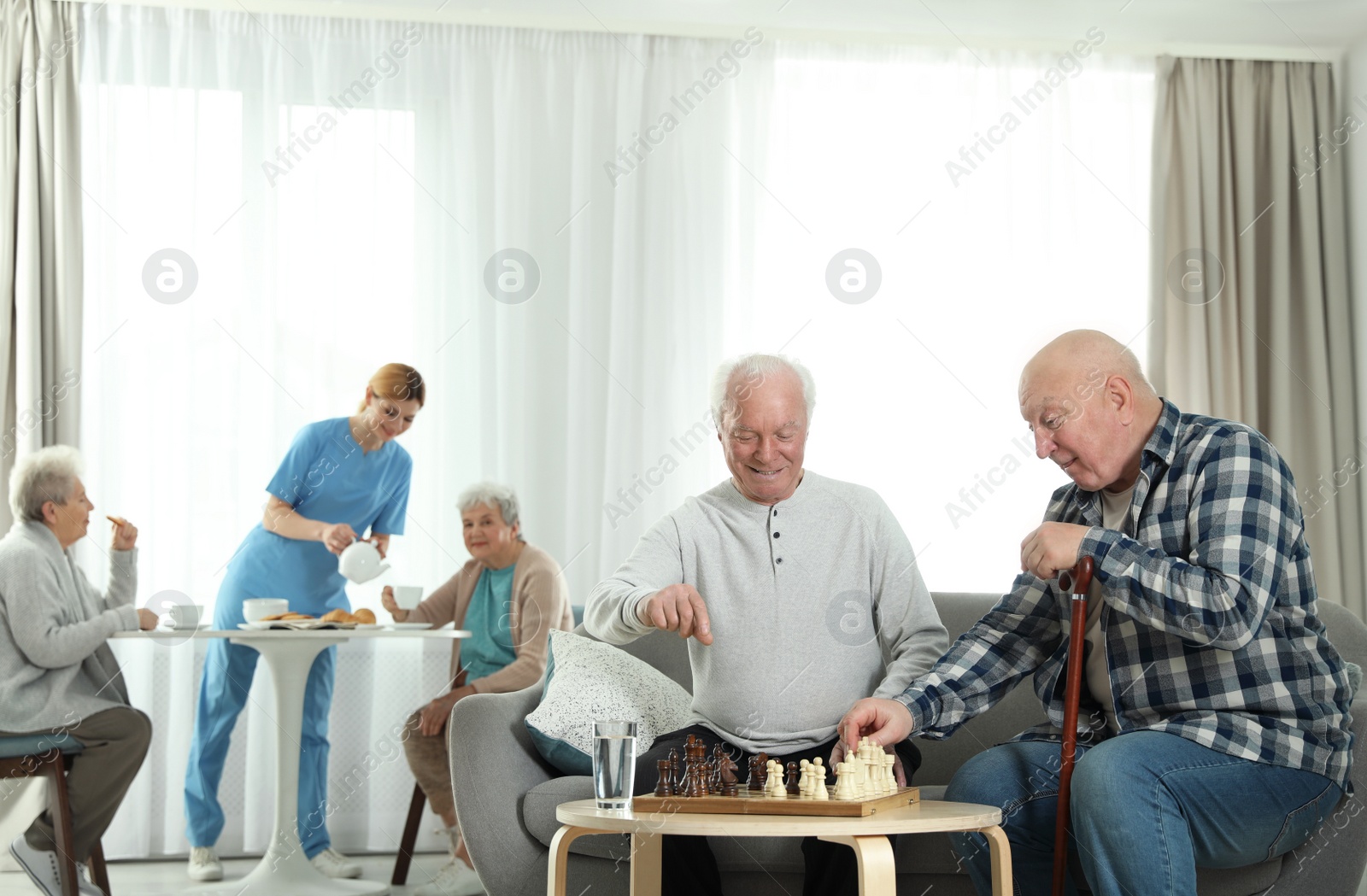 Photo of Elderly men playing chess while nurse serving breakfast to women at retirement home. Assisting senior people