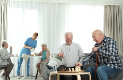 Photo of Elderly men playing chess while nurse serving breakfast to women at retirement home. Assisting senior people
