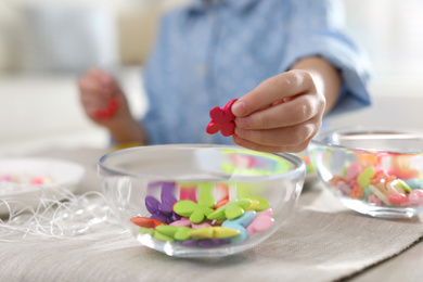 Photo of Little girl making accessory with beads at table indoors, closeup. Creative hobby