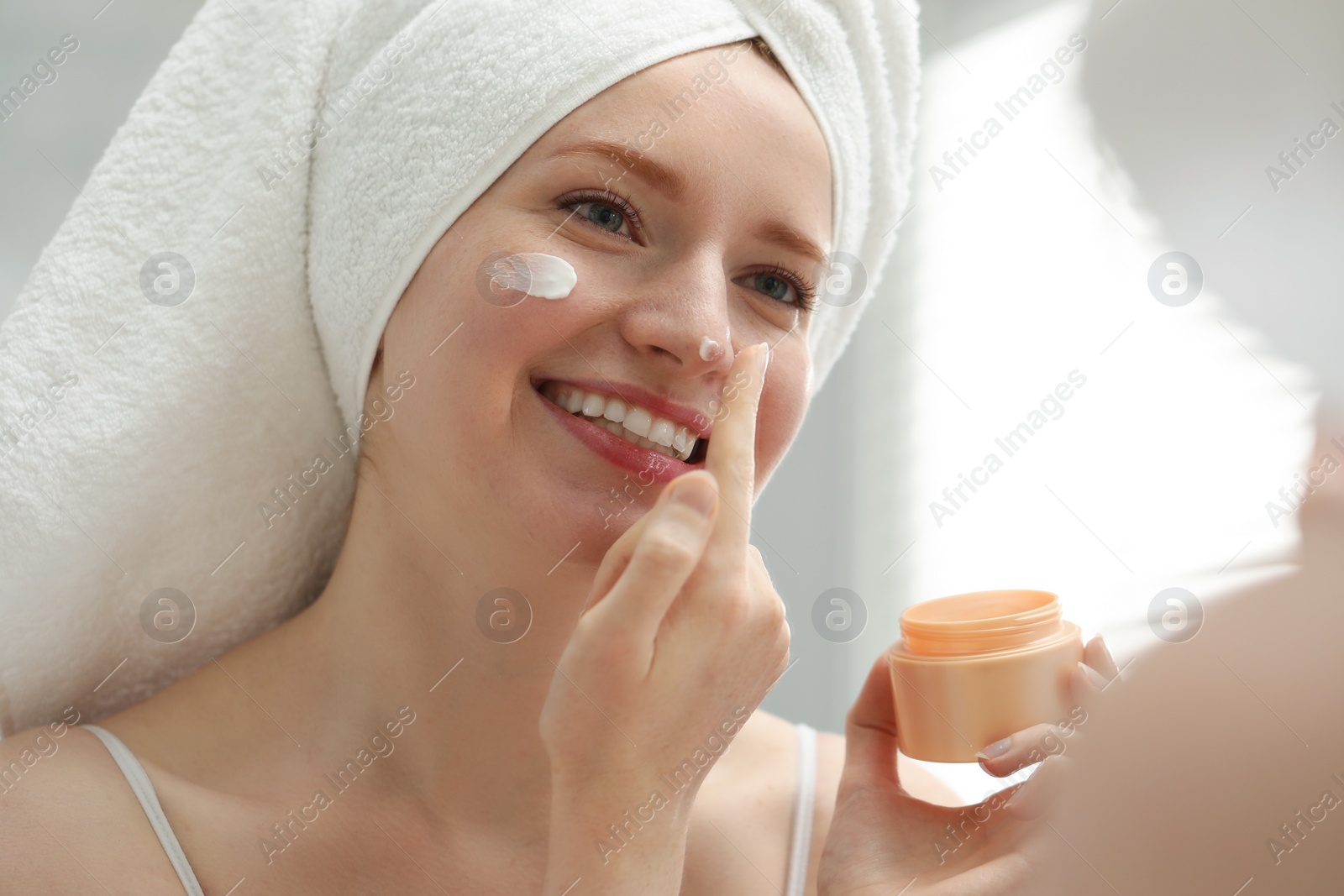 Photo of Smiling woman with freckles applying cream onto her face in bathroom