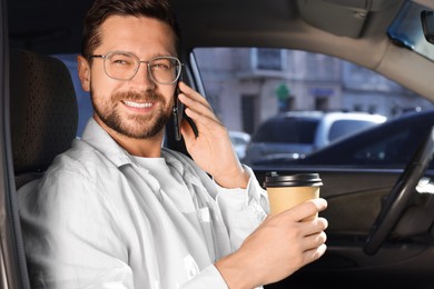 Photo of Coffee to go. Happy man with paper cup of drink talking on smartphone in car
