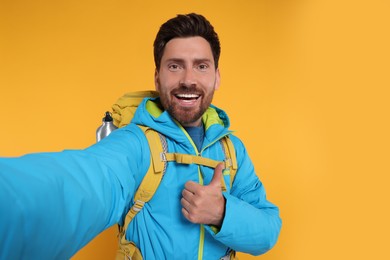 Photo of Happy man with backpack taking selfie and showing thumb up on orange background. Active tourism
