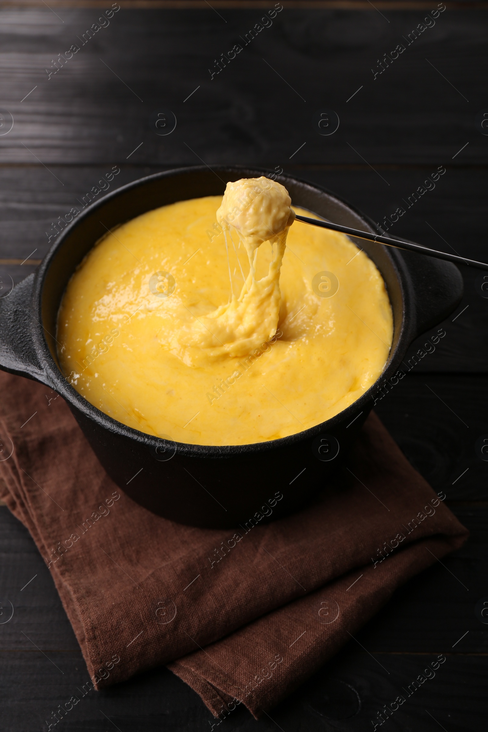 Photo of Dipping piece of ham into fondue pot with melted cheese on black wooden table