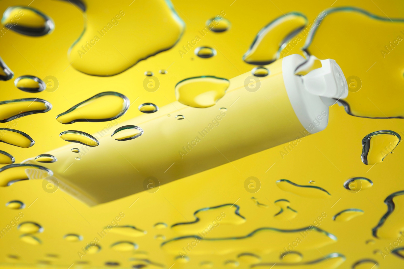 Photo of Moisturizing cream in tube on glass with water drops against yellow background