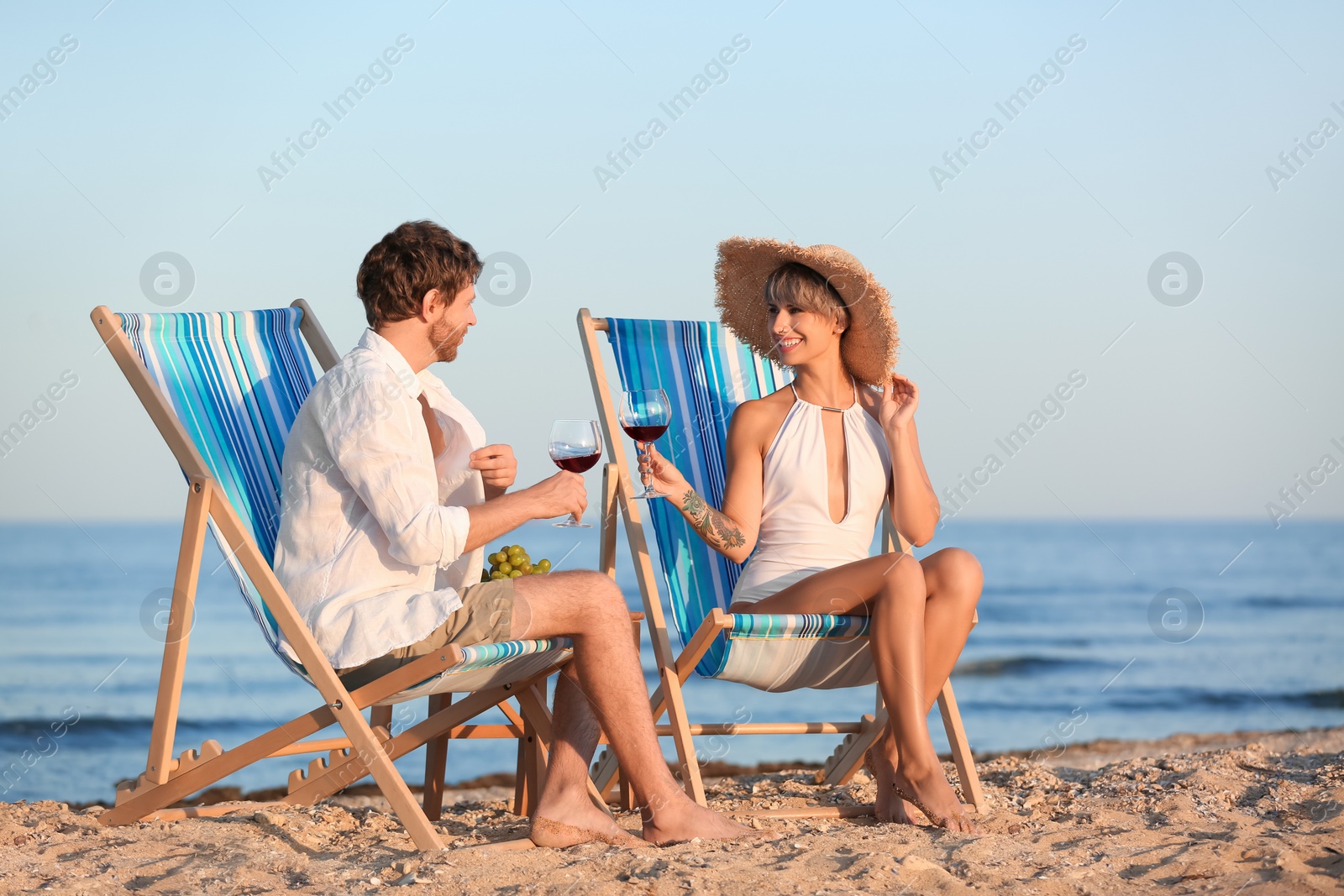 Photo of Young couple with glasses of wine in beach chairs at seacoast