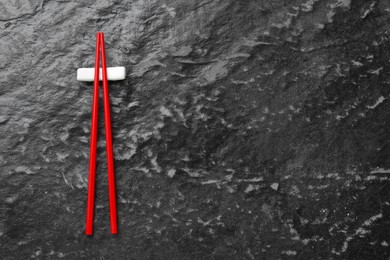 Photo of Pair of red chopsticks with rest on black textured table, top view. Space for text