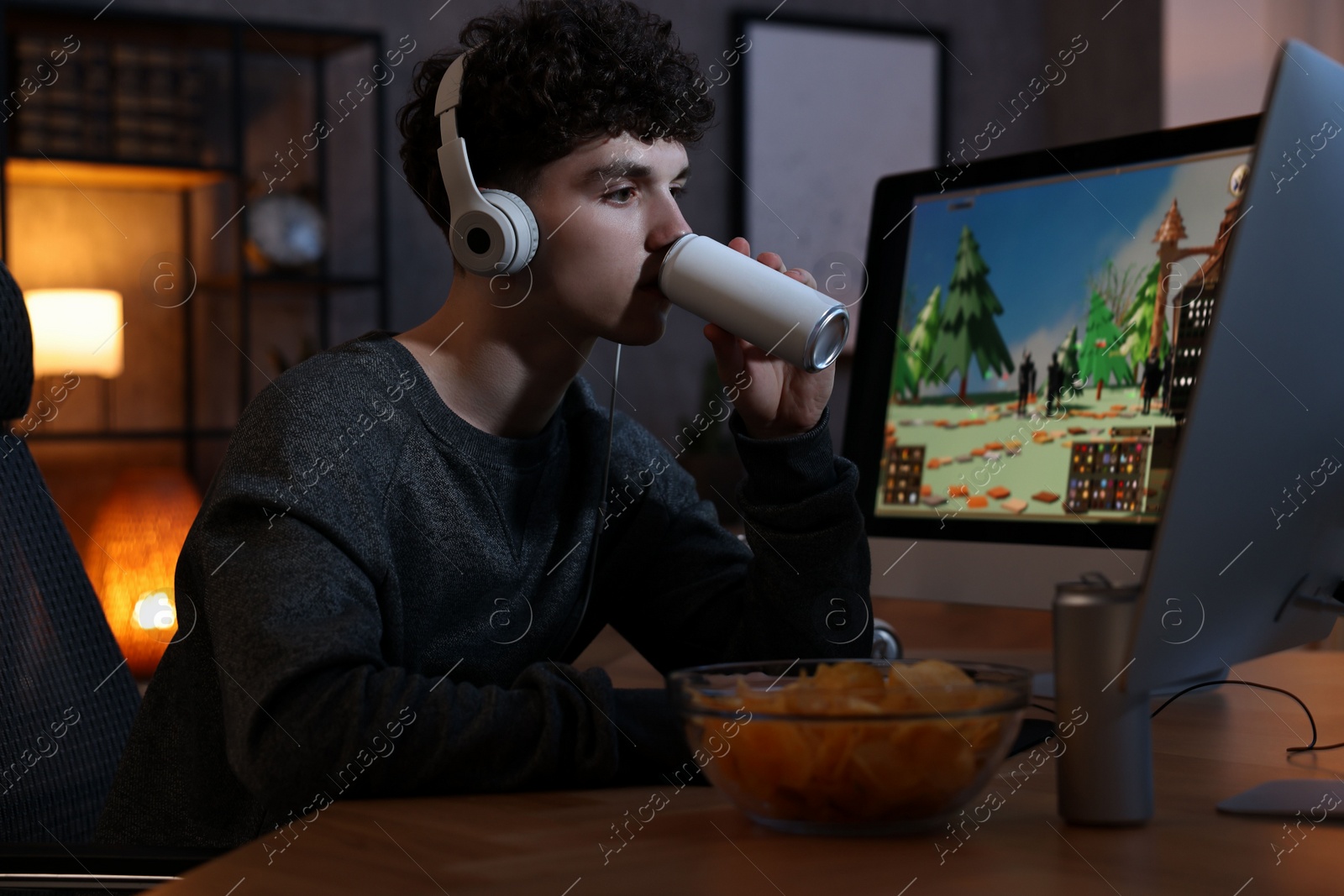 Photo of Young man with energy drink and headphones playing video game at wooden desk indoors