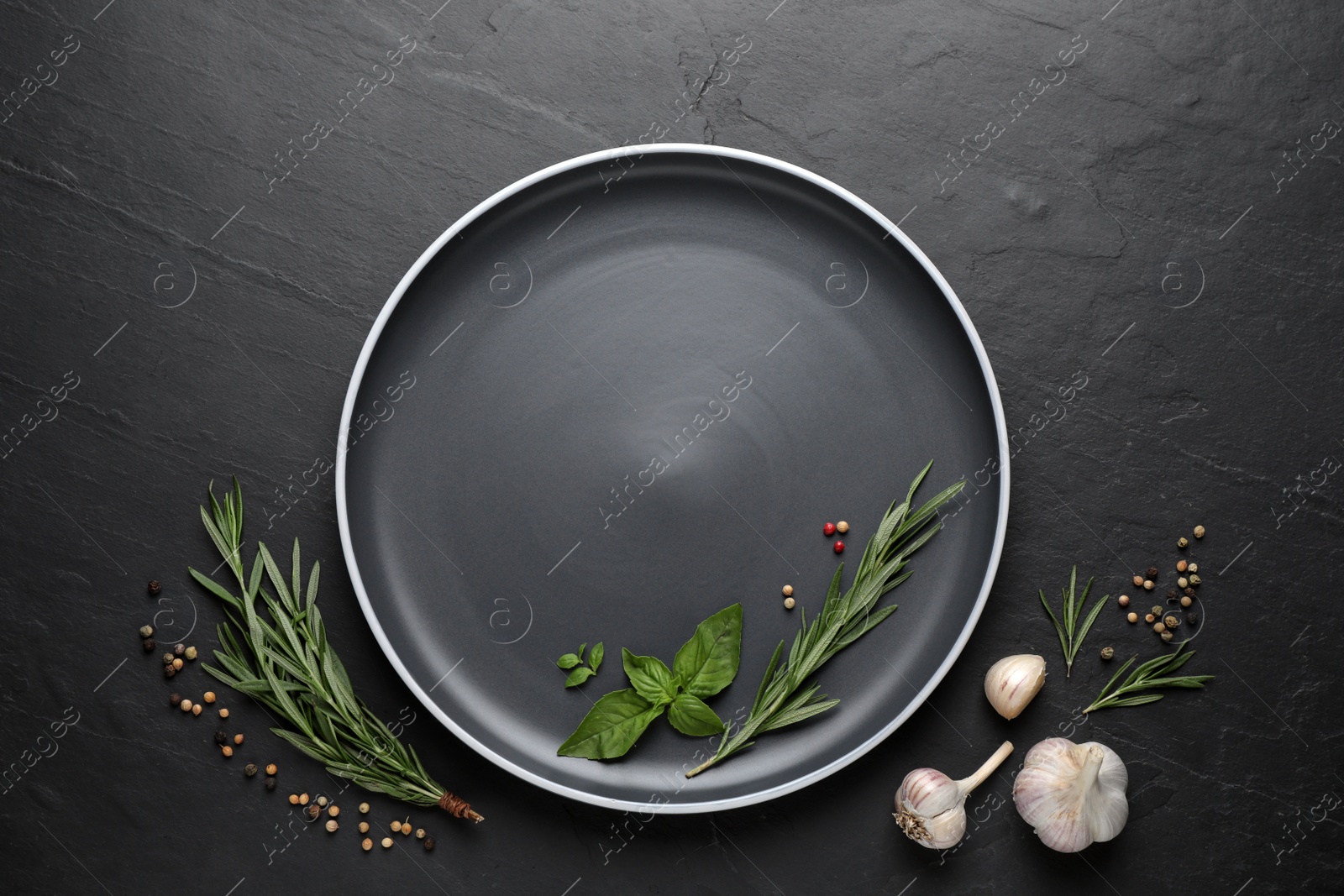 Photo of Dark plate with herbs, peppercorns and garlic on black table, flat lay