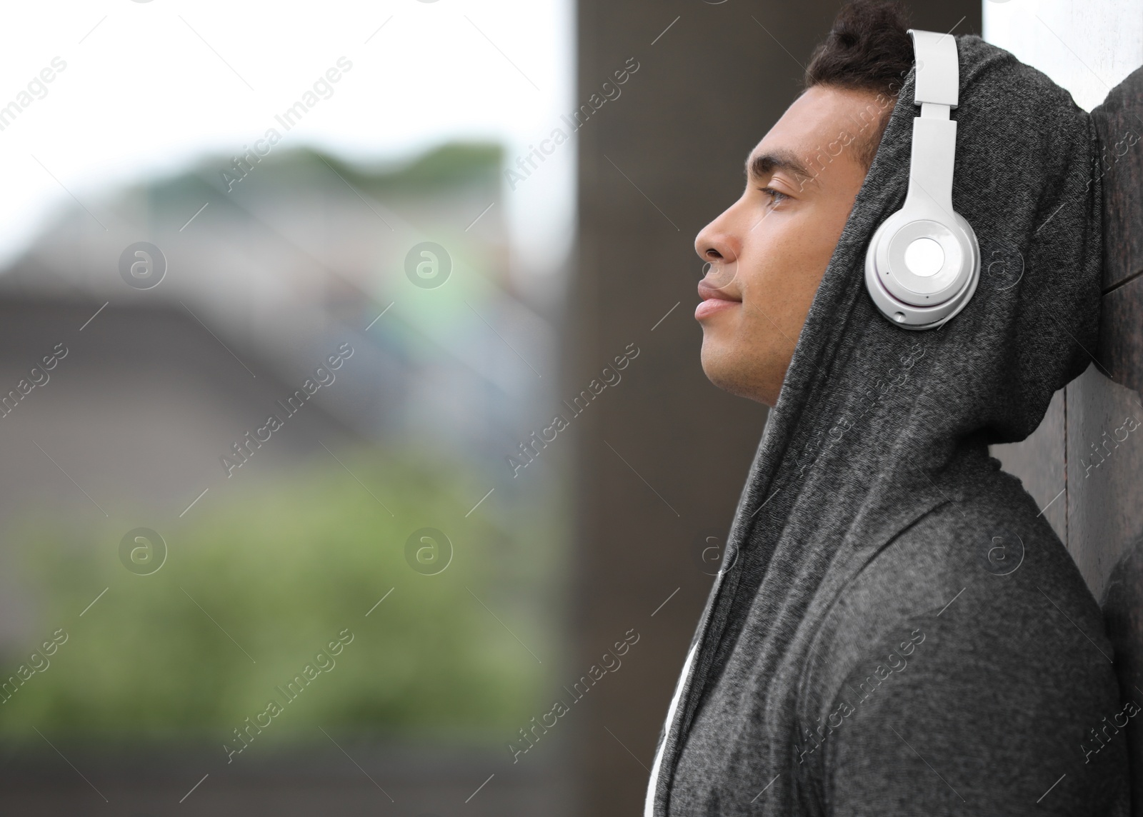 Photo of Handsome young African-American man with headphones listening to music near wall on city street. Space for text
