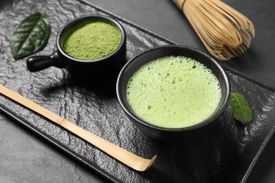 Photo of Cup of fresh matcha tea, bamboo whisk, spoon and green powder on black table, closeup