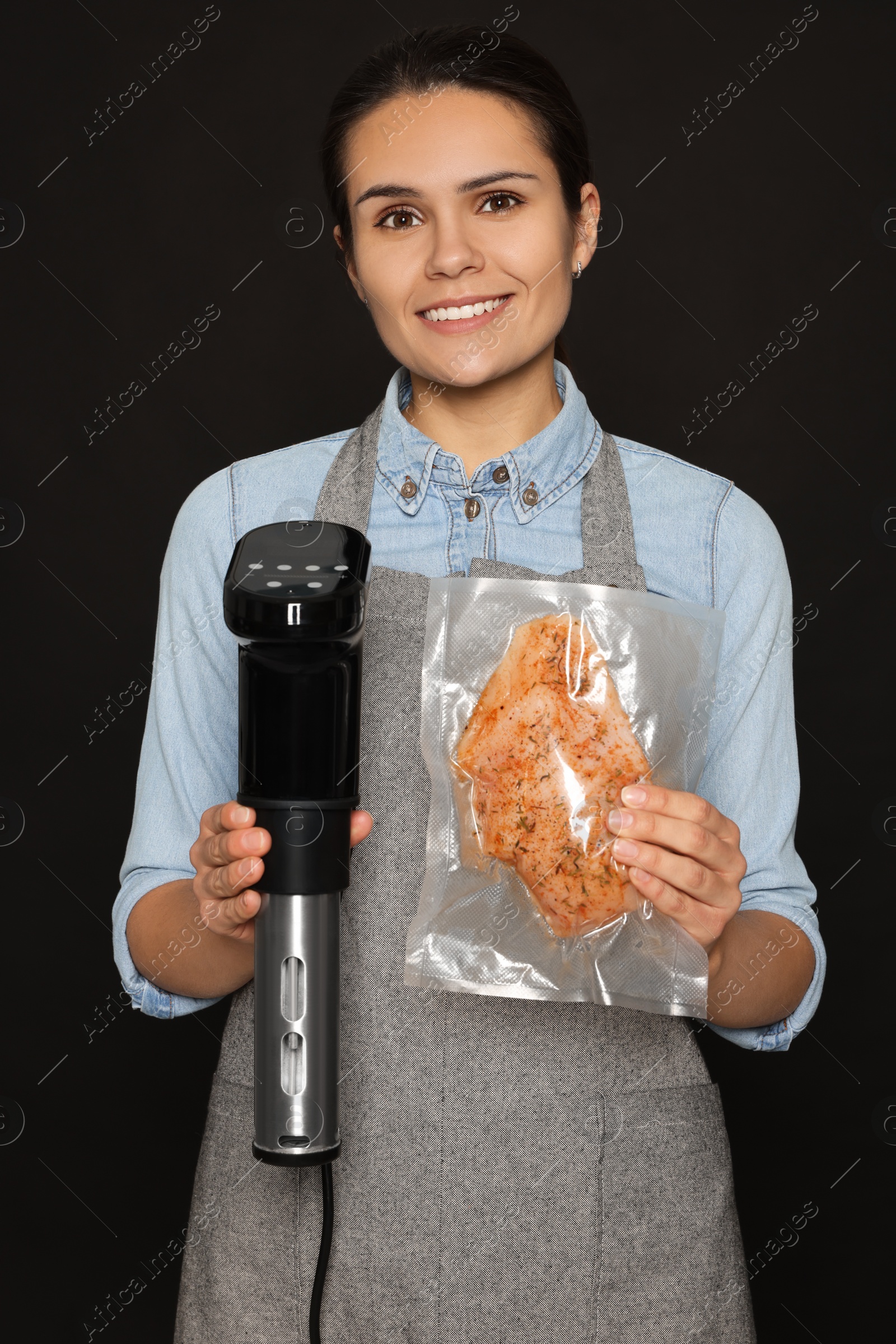 Photo of Beautiful young woman holding sous vide cooker and meat in vacuum pack on black background