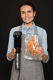 Photo of Beautiful young woman holding sous vide cooker and meat in vacuum pack on black background