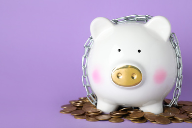 Photo of Piggy bank  with steel chain and coins and lilac background, space for text. Money safety concept