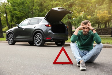 Photo of Upset man sitting near warning triangle and broken car on road