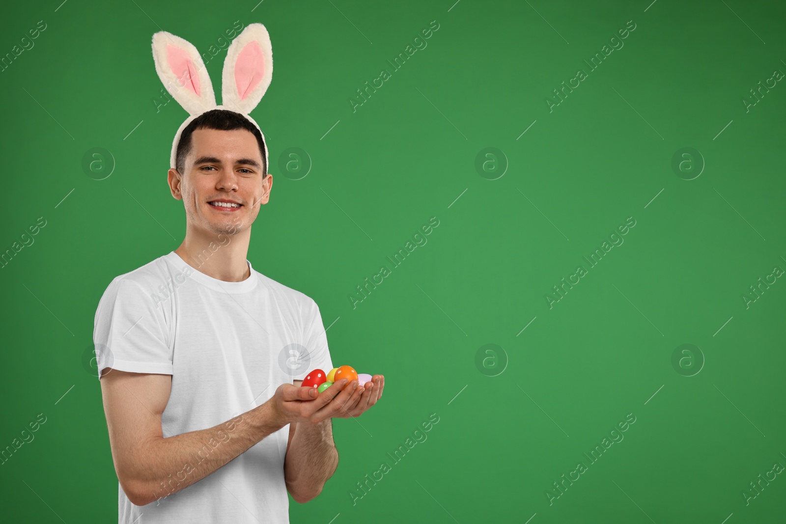 Photo of Easter celebration. Handsome young man with bunny ears holding painted eggs on green background. Space for text