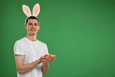 Easter celebration. Handsome young man with bunny ears holding painted eggs on green background. Space for text