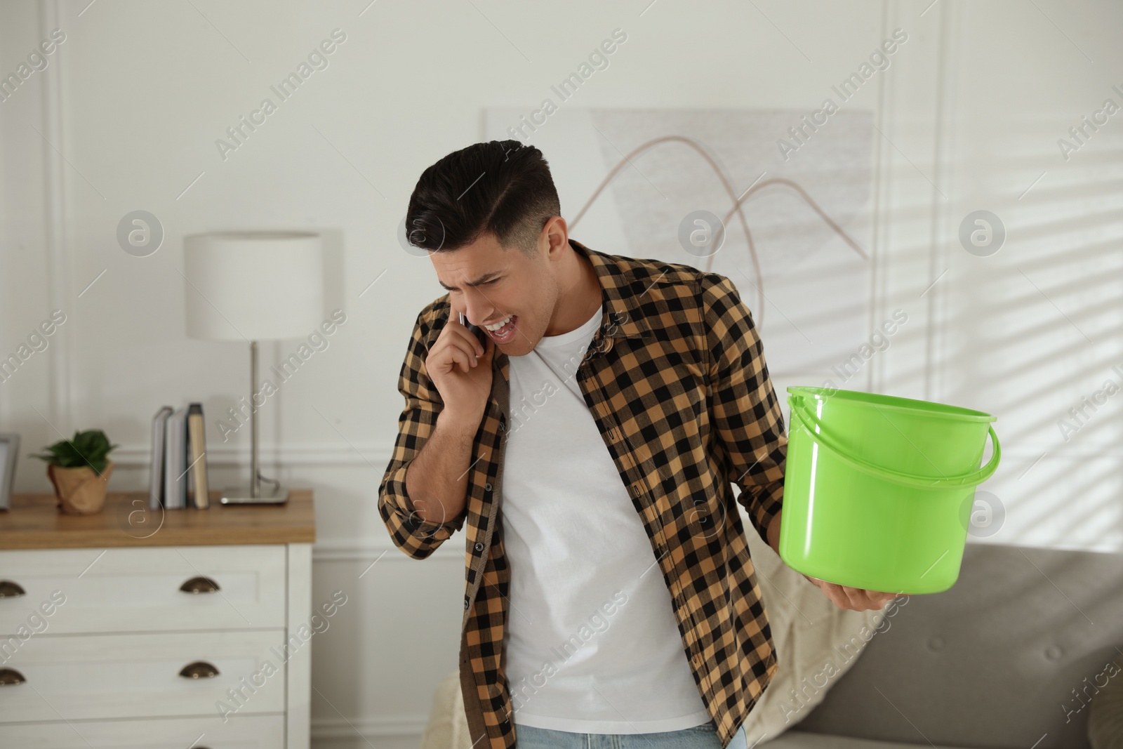 Photo of Emotional man calling roof repair service while collecting leaking water from ceiling in living room