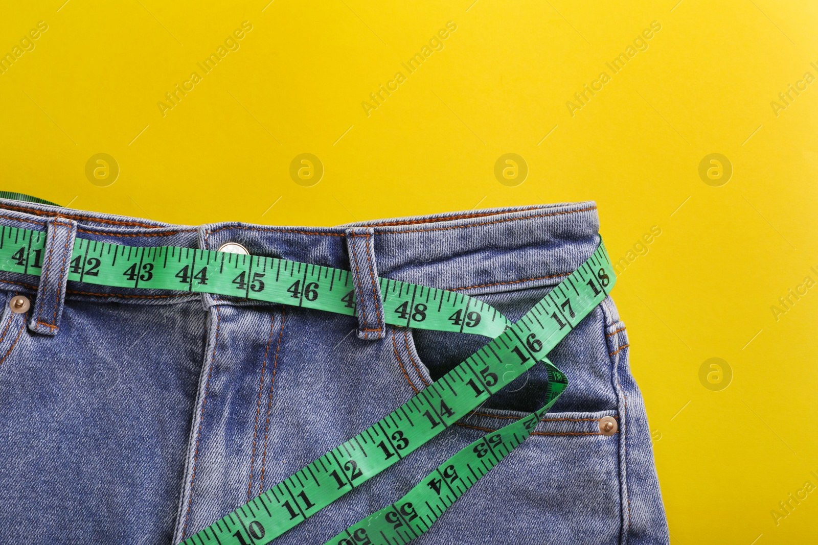 Photo of Jeans with measuring tape on yellow background, top view. Weight loss concept