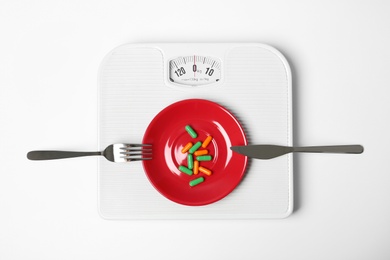 Photo of Scales with plate of weight loss pills and cutlery on white background, top view