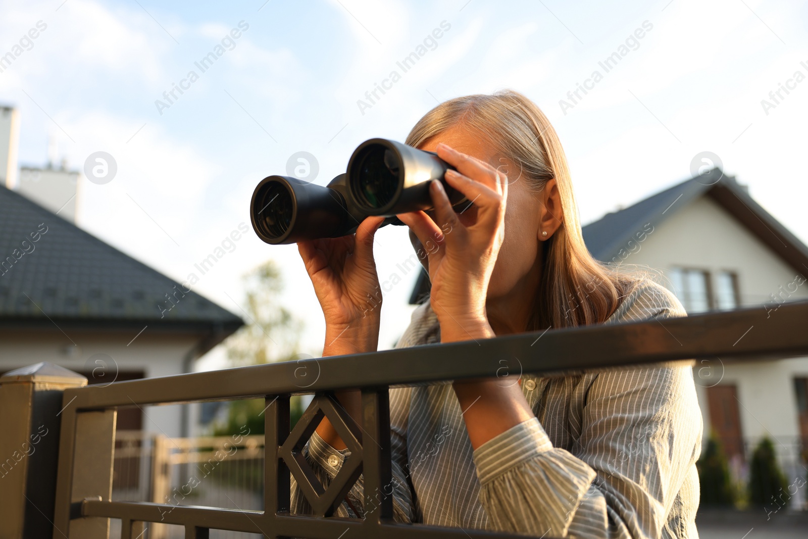 Photo of Concept of private life. Curious senior woman with binoculars spying on neighbours over fence outdoors