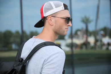 Photo of Handsome young man with stylish sunglasses and backpack near reflection surface outdoors