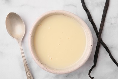 Photo of Bowl with condensed milk, vanilla pods and spoon on white marble table, flat lay
