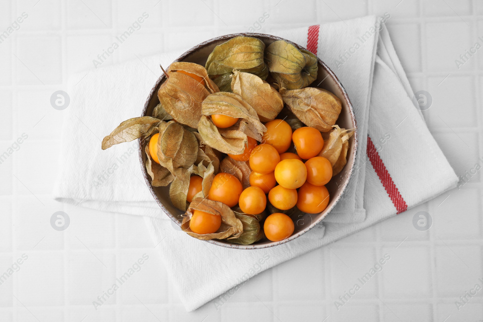 Photo of Ripe physalis fruits with calyxes in bowl on white tiled table, top view