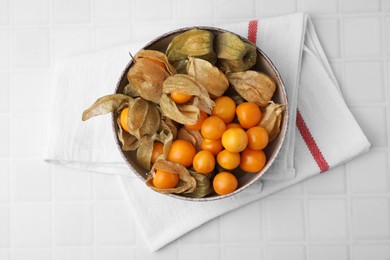 Ripe physalis fruits with calyxes in bowl on white tiled table, top view