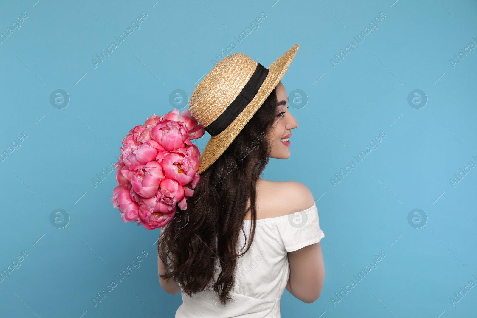 Photo of Beautiful young woman in straw hat with bouquet of pink peonies against light blue background